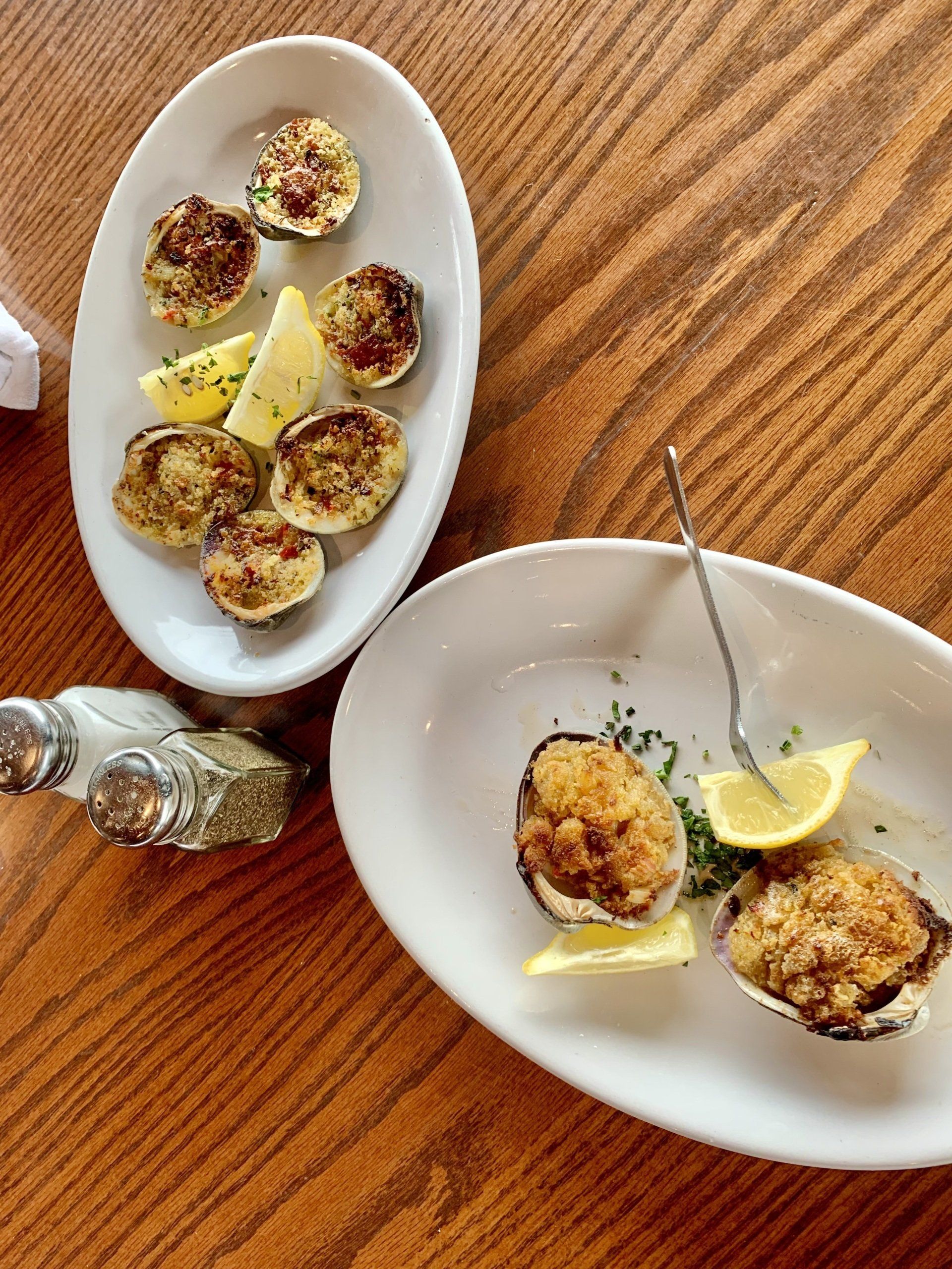 Two white plates holding baked oysters with breadcrumbs and lemon wedges on a wooden table, with salt and pepper shakers.