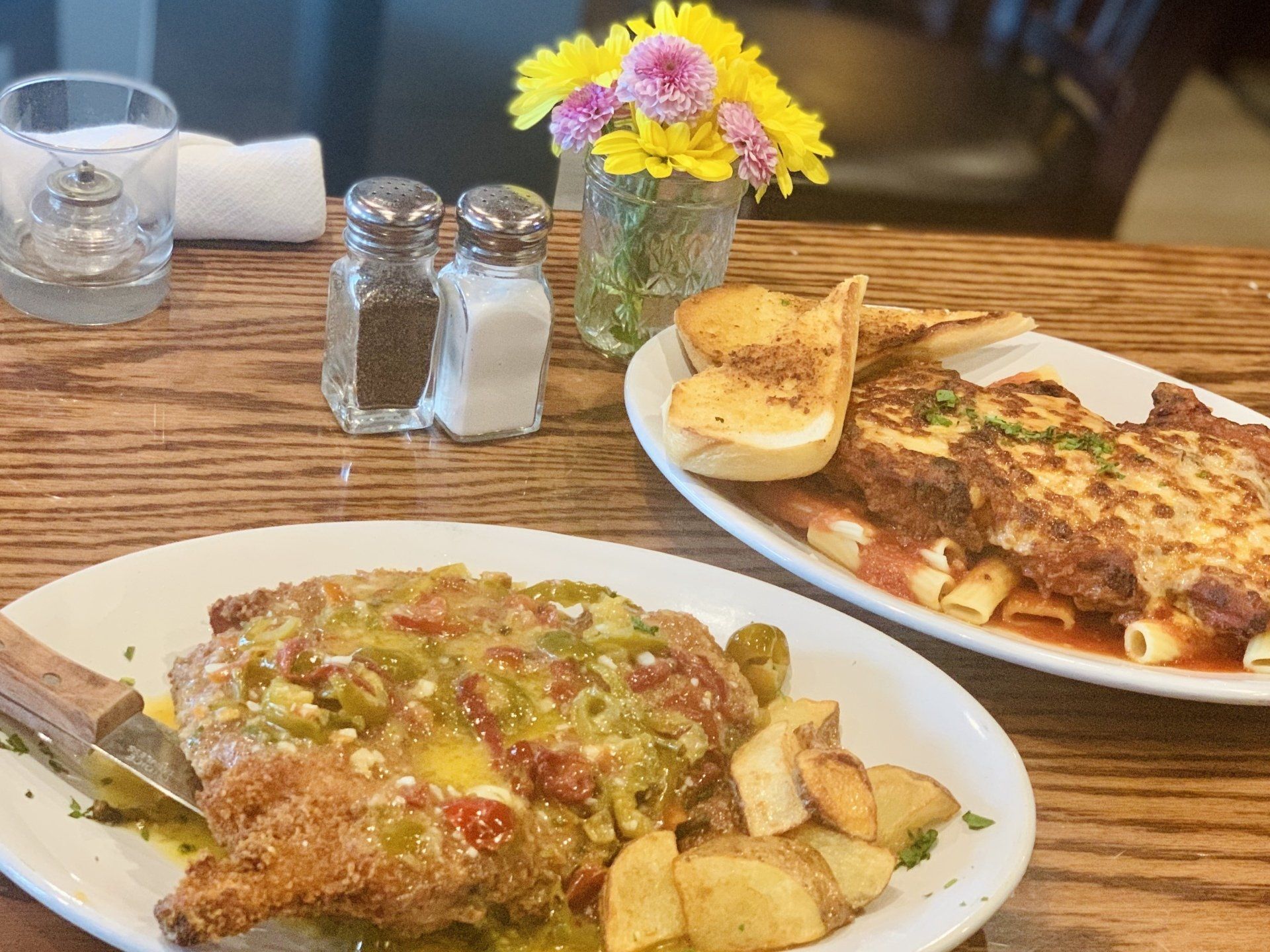 Two plates of breaded, sauce-topped entrees with sides sit on a wooden table near salt and pepper shakers and flowers.