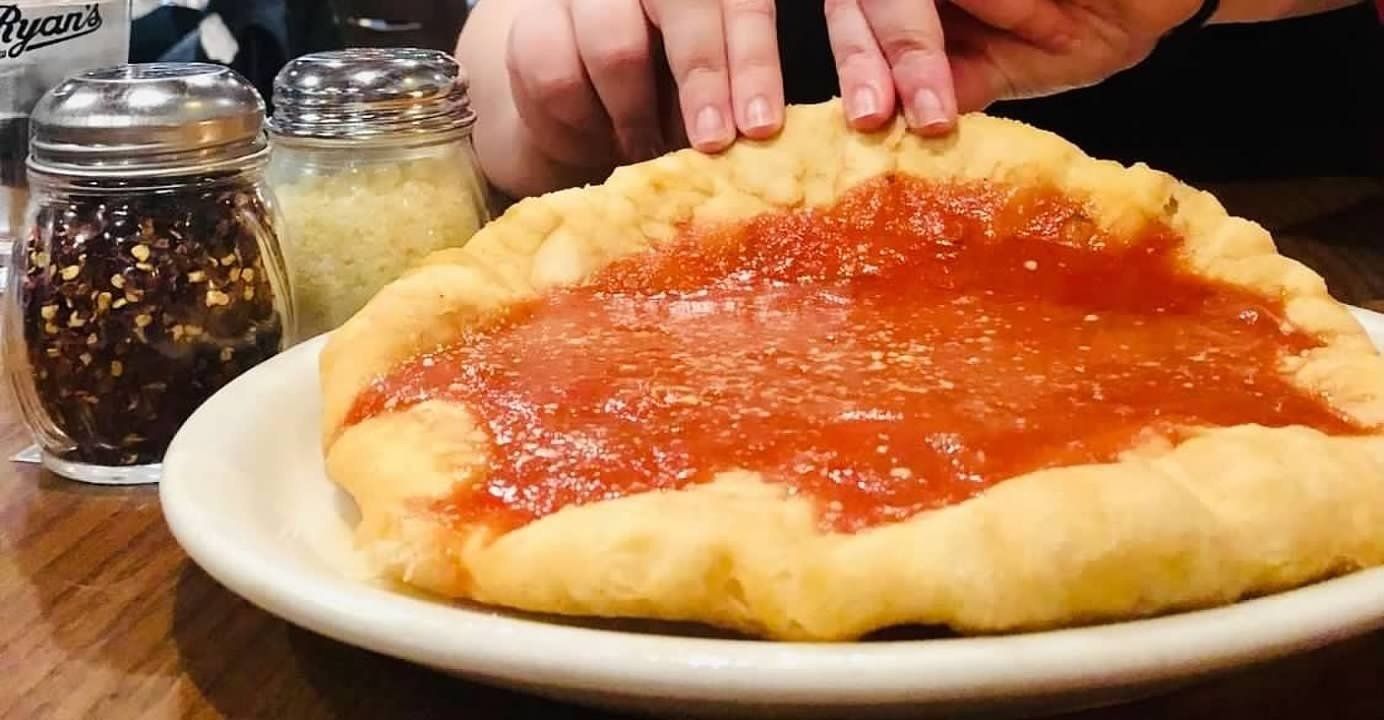 A person's hands holding the crust of a round, deep-fried pizza with red sauce on a plate, with shakers on the table.