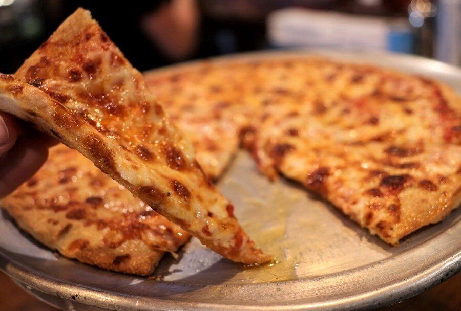 A hand holds a slice of cheese pizza above a metal tray containing the rest of the pie.