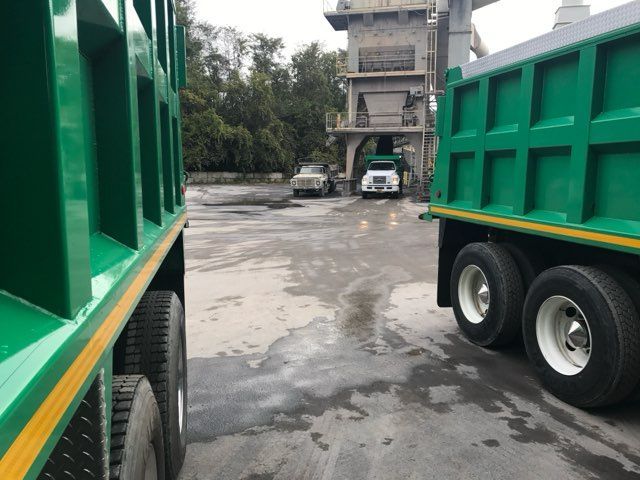 Green dump trucks parked on a wet lot beside a building, with trees in the background.