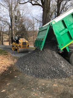 Green dump truck unloading black gravel near a yellow skid steer on a tree-lined road.