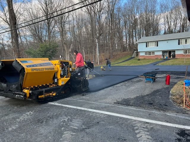 Road paving machine laying black asphalt while a worker in red stands nearby on a residential street