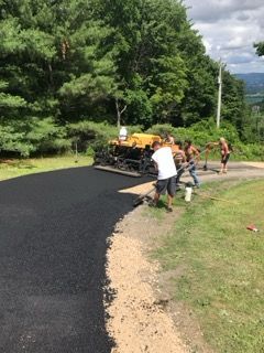 Workers paving a curved road with fresh black asphalt beside a grassy area and trees