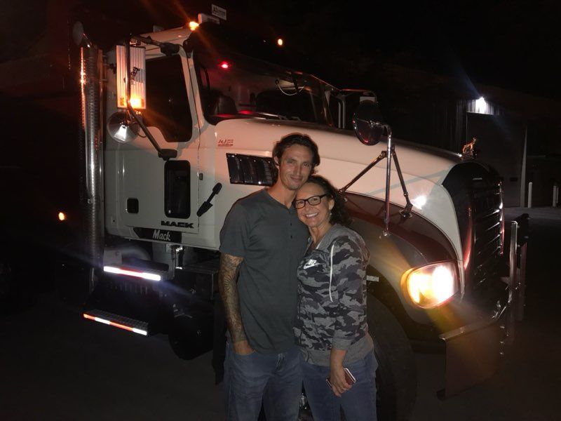 Two people posing in front of a white semi-truck at night, one smiling beside the lit headlight.