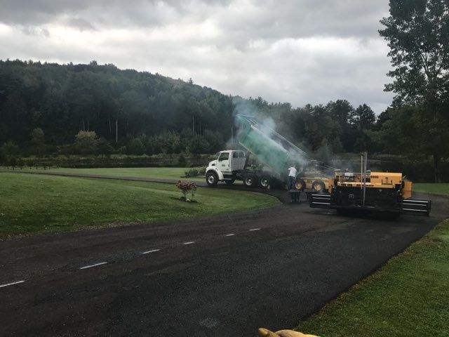 Road paving machine and truck smoking on a rural roadside under cloudy skies