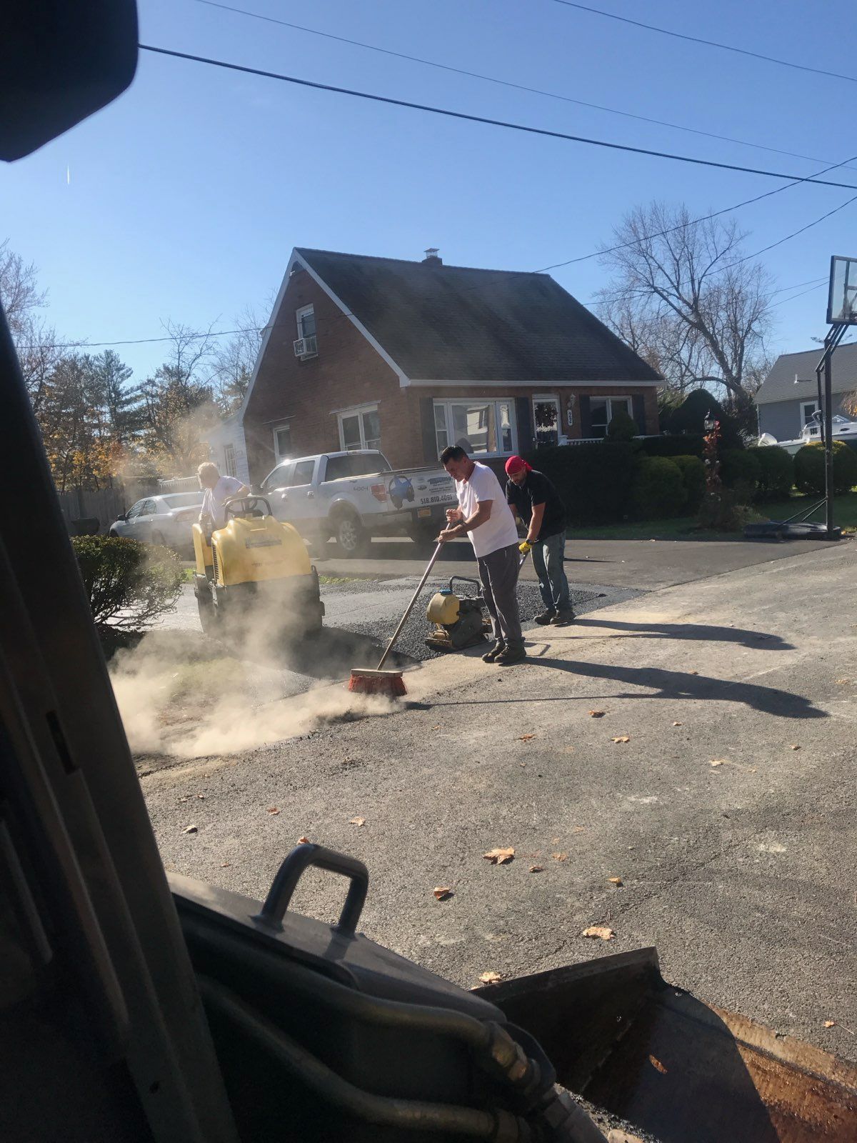 People clearing debris on a sunny residential street, with smoke or dust rising near a brick house.