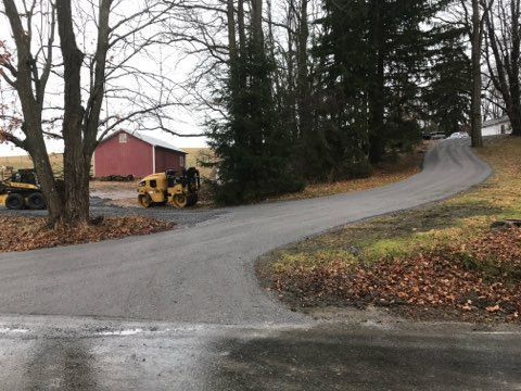 Curving gravel driveway beside bare trees, a red barn, and a yellow tractor on a cloudy day