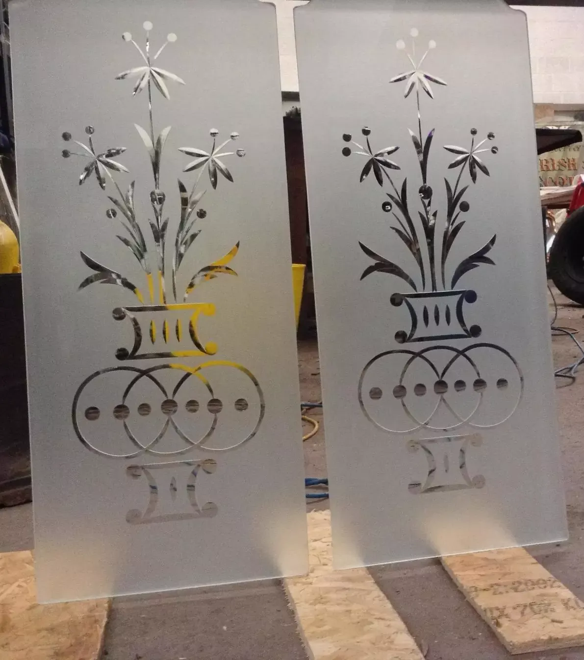 Two frosted glass panels with etched vase and flower designs in a workshop setting