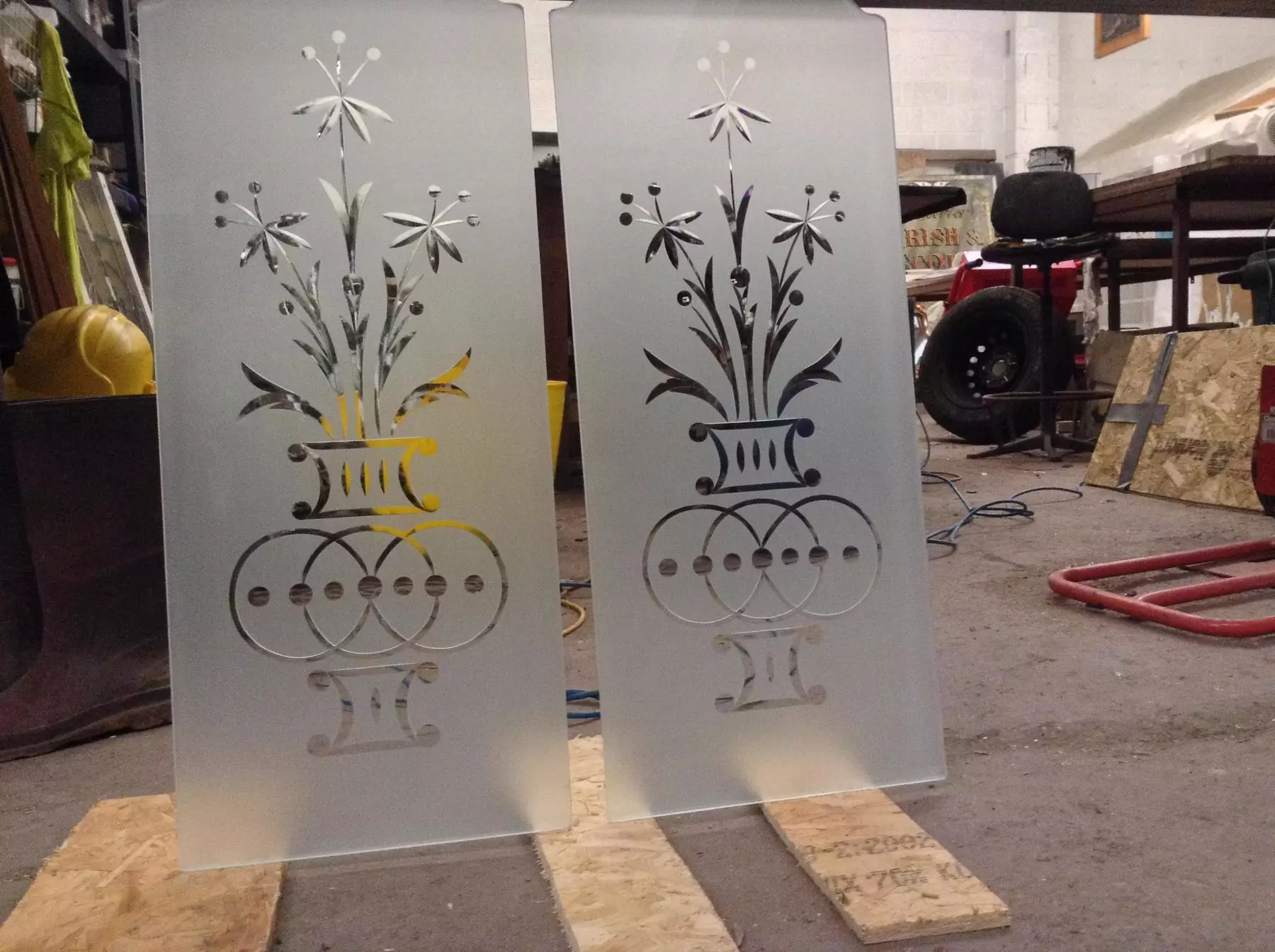 Two frosted glass panels with black floral and geometric designs standing in a workshop.