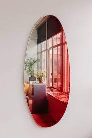 Oval wall mirror reflecting a red-tinted modern room with large windows and plants
