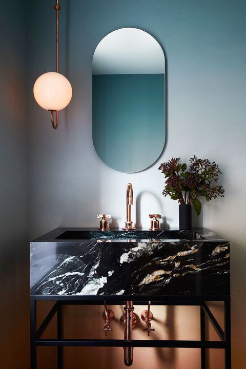 Modern bathroom vanity with black marble sink, oval mirror, brass fixtures, and wall sconce on pale blue wall