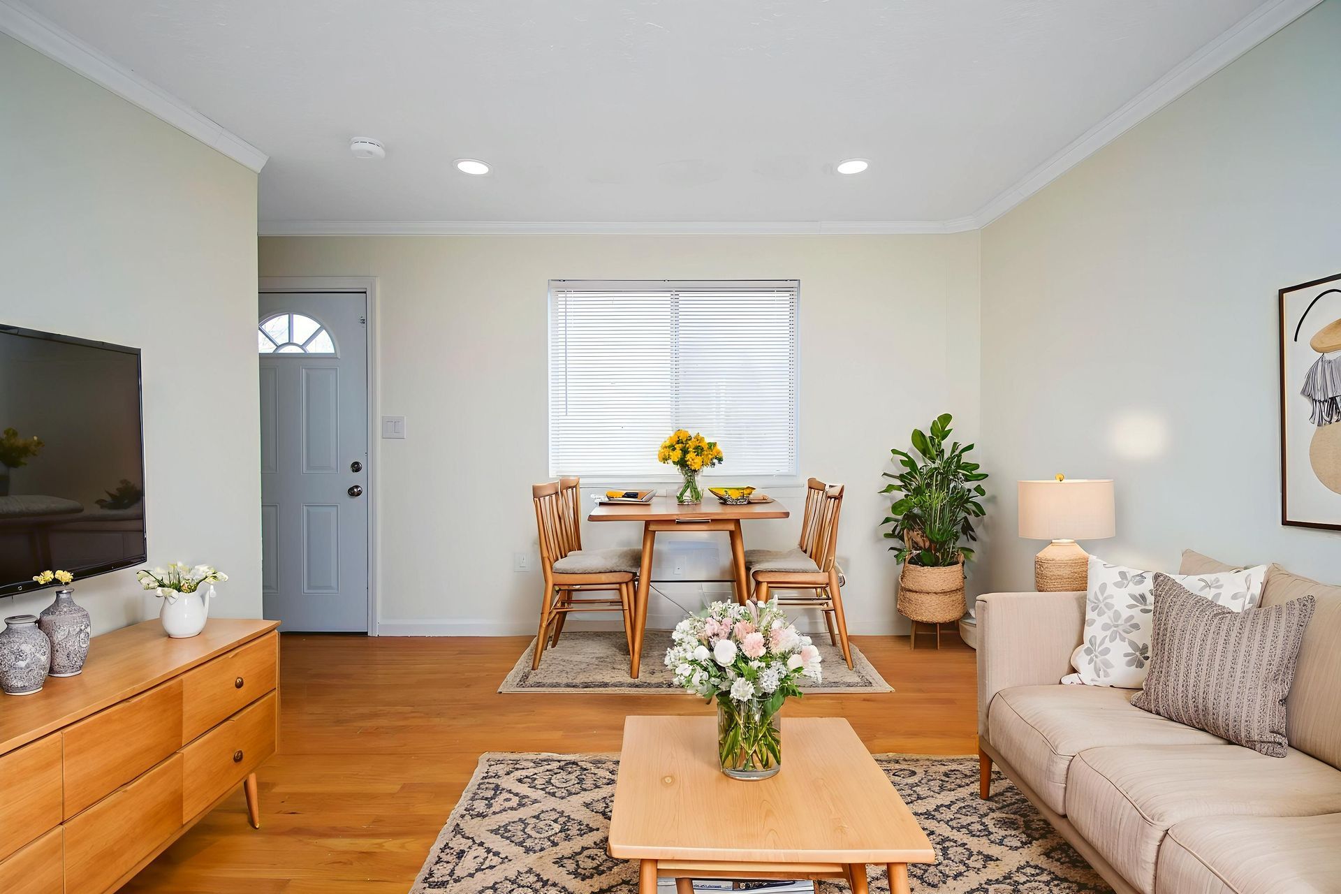 Bright living room with beige sofa, wooden coffee table, dining nook, and potted plants.