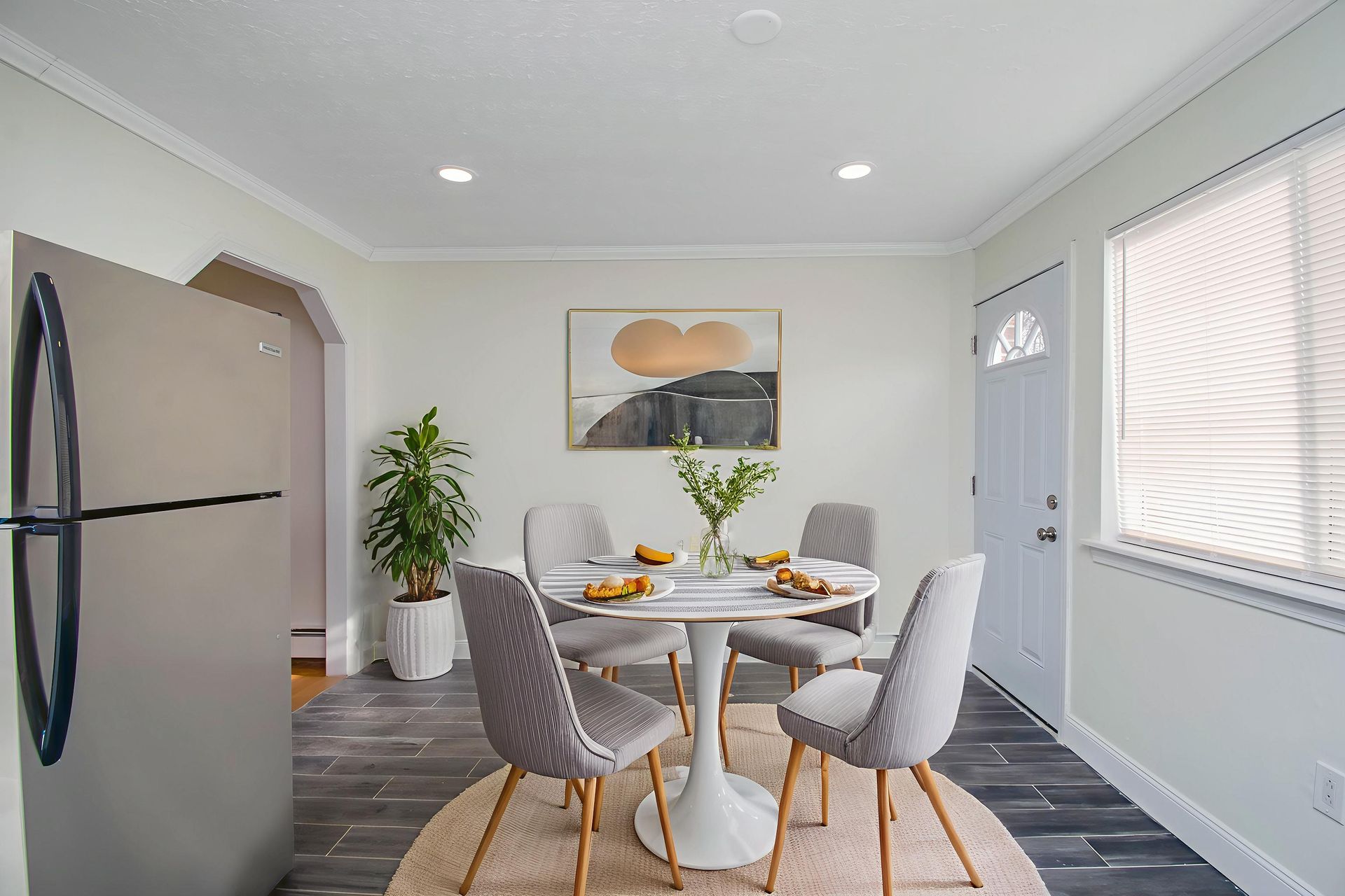 Modern dining room with round table, gray chairs, and a kitchen doorway beside a window.