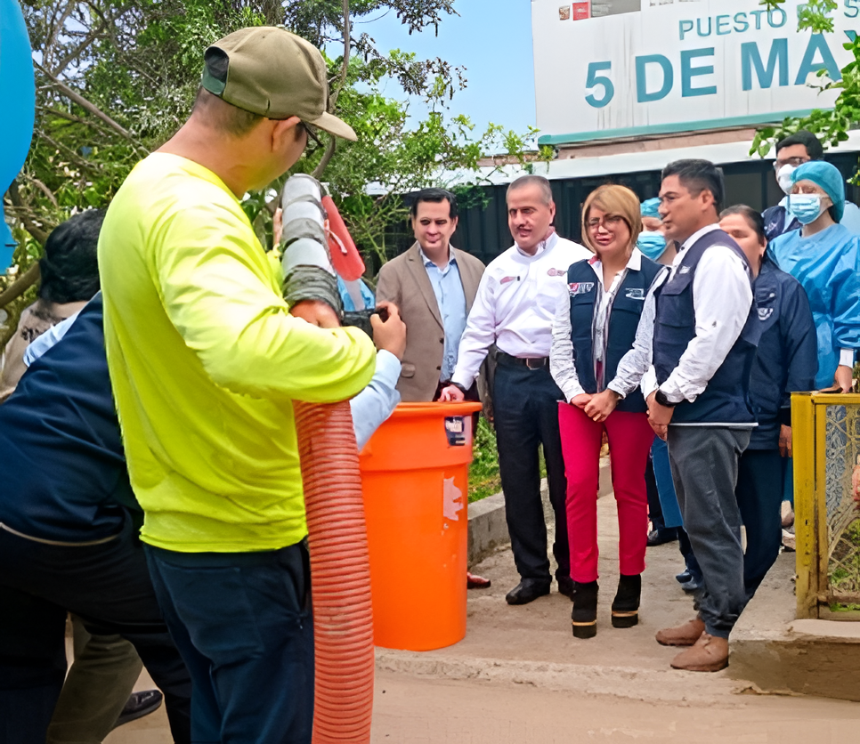 Varias personas se congregaron al aire libre alrededor de un barril naranja; un hombre con una camisa de color neón habla con los funcionarios cerca de un letrero.