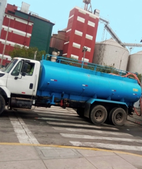 Camión cisterna azul estacionado en una calle junto a edificios industriales rojos y silos.