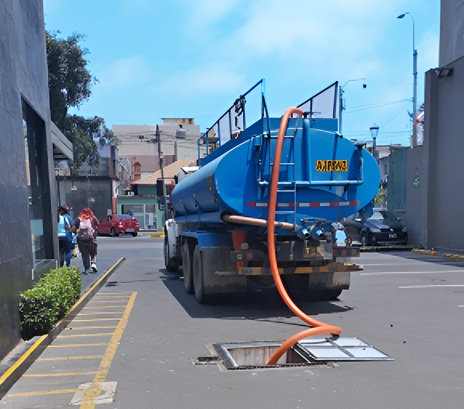 Camión cisterna azul estacionado en la calle con una manguera naranja conectada a una alcantarilla abierta.