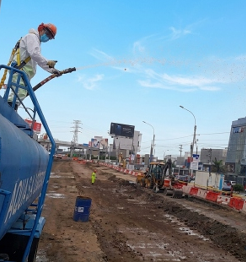 Obrero de la construcción en un camión azul rociando con agua lodosa la carretera en una zona de obras viales bajo un cielo azul.