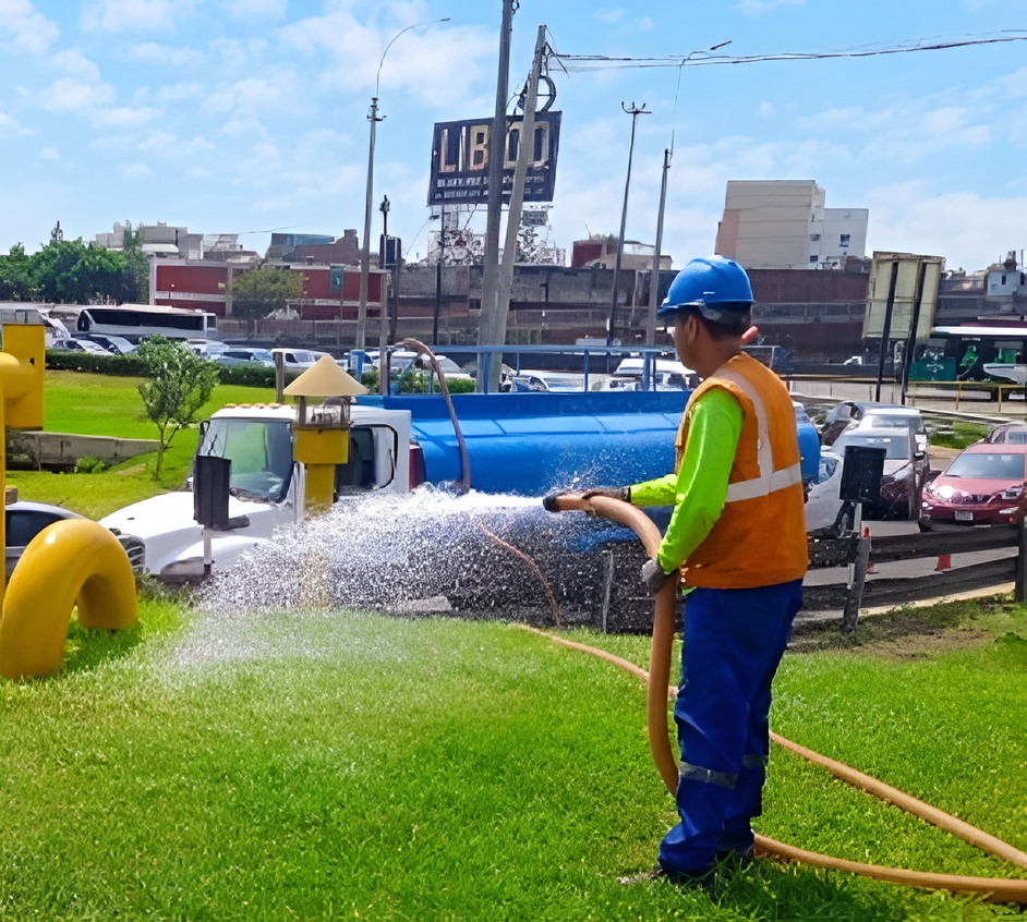 Un trabajador con chaleco naranja y casco azul rocía agua sobre el césped verde al borde de la carretera.