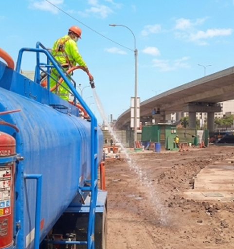 Un trabajador con ropa reflectante rocía agua desde un camión cisterna azul en una obra de construcción de una carretera embarrada.