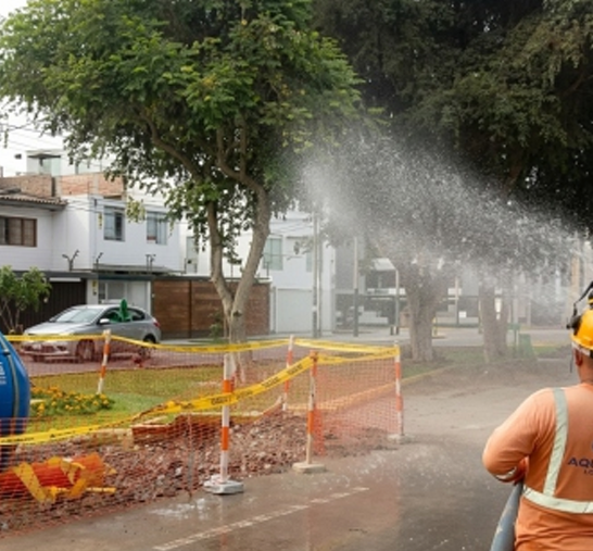 Un trabajador rocía agua en una calle junto a una valla de seguridad naranja y árboles en un barrio de la ciudad.
