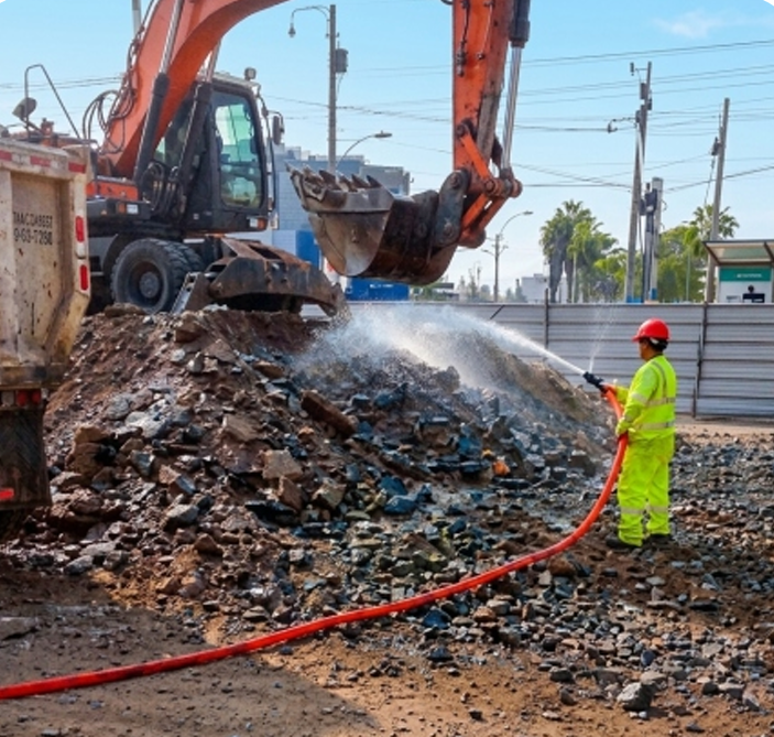 Un trabajador con un traje de seguridad de color neón rocía agua sobre los escombros junto a una excavadora en una obra de demolición.