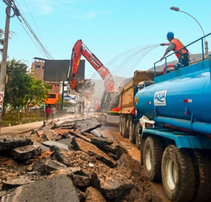 Una excavadora y un camión cisterna trabajan para retirar los escombros de una carretera dañada, mientras el agua se rocía sobre los restos.