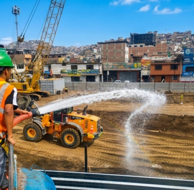 Un obrero de la construcción rocía agua sobre un terreno baldío junto a un camión volquete naranja y una grúa en una obra.