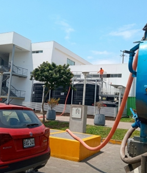 Coche rojo aparcado junto a un edificio blanco con un trabajador en el tejado y mangueras naranjas en primer plano.