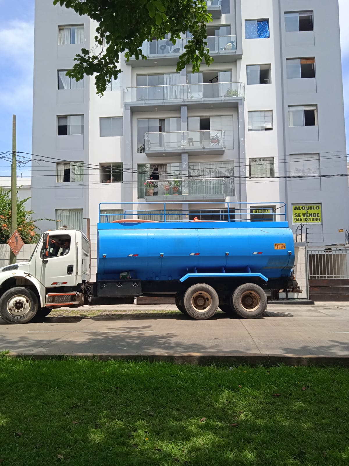 Un camión cisterna azul estacionado frente a un edificio de apartamentos blanco, con un césped verde en primer plano.