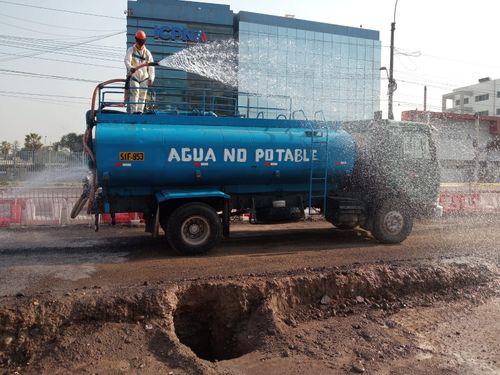 Un camión cisterna azul rocía agua sobre una calle fangosa cerca de edificios, con la inscripción