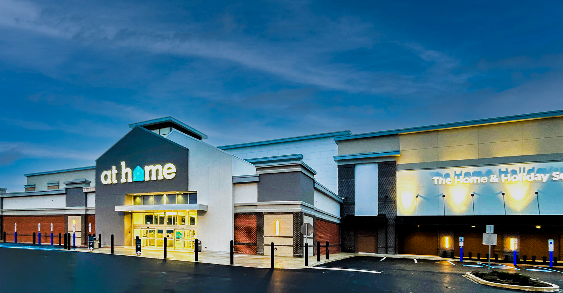 At Home store exterior at dusk, with logo, entrance, and parking lot.