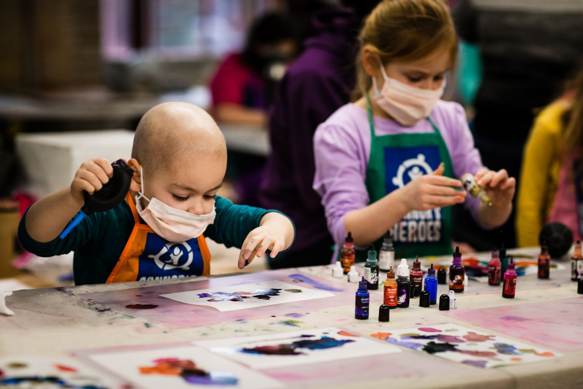 Children in masks create art at a table; one child wears an apron.