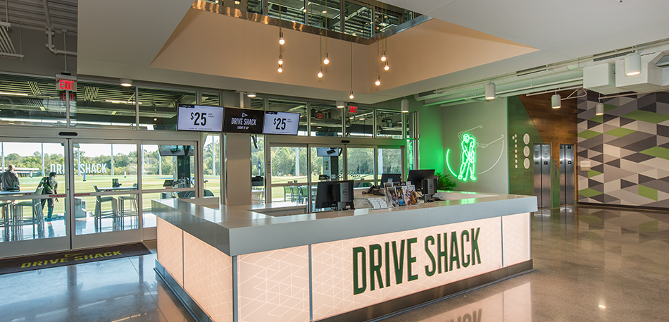 Drive Shack front desk. A modern interior with a white counter and green logo, overlooking a golf course.