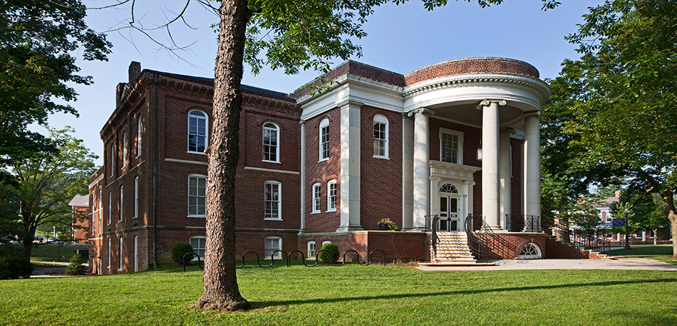 Brick building with a white columned entrance. Green lawn and trees surround the structure.