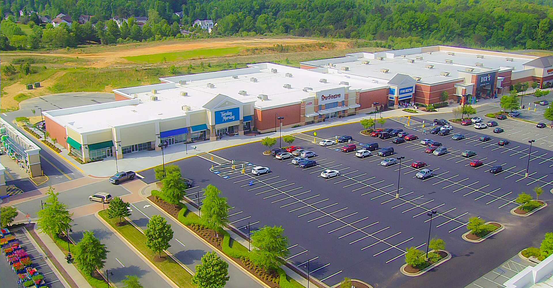 Aerial view of a large shopping center with a vast parking lot. Buildings have white and tan facades.