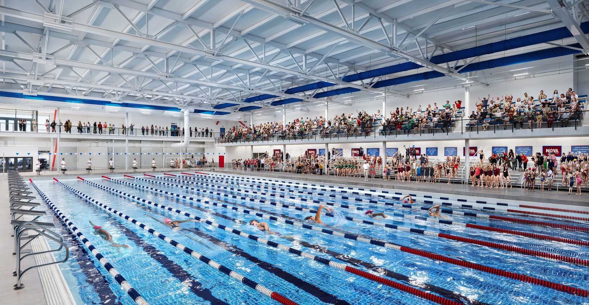 Indoor Olympic-size swimming pool with lane lines and spectators in bleachers, brightly lit under a blue roof
