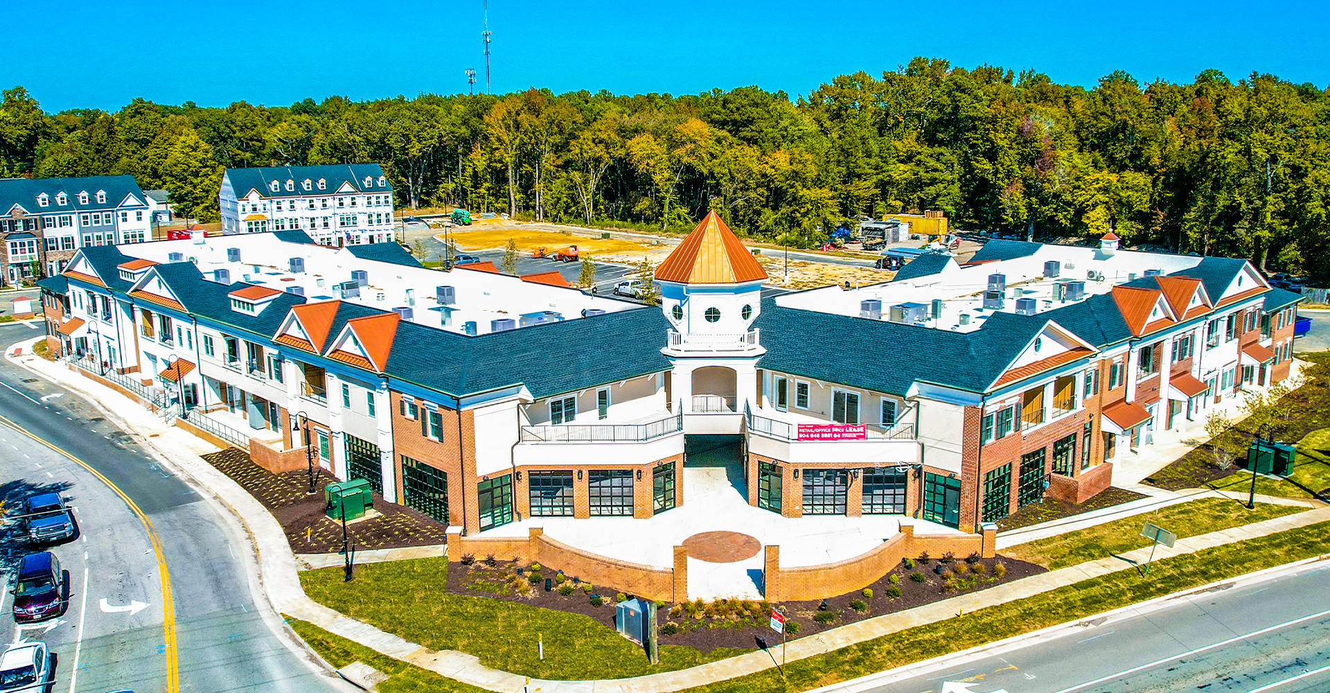 Aerial view of a complex with brick buildings, dark roofs, and a central tower, with trees in the background.