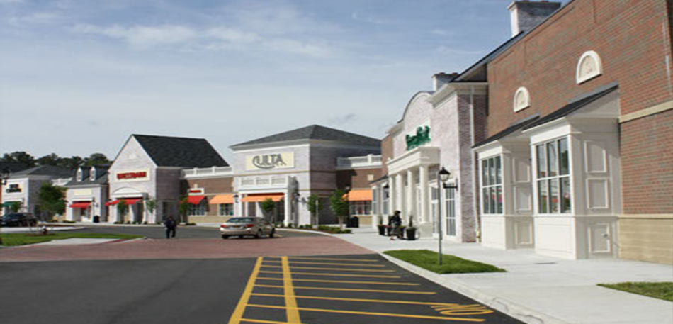 Shopping center with brick and light-colored buildings, parked cars, and a blue sky.