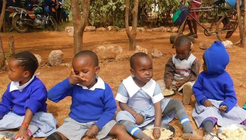 Children in blue school uniforms eating outside. Brown ground, trees in background.