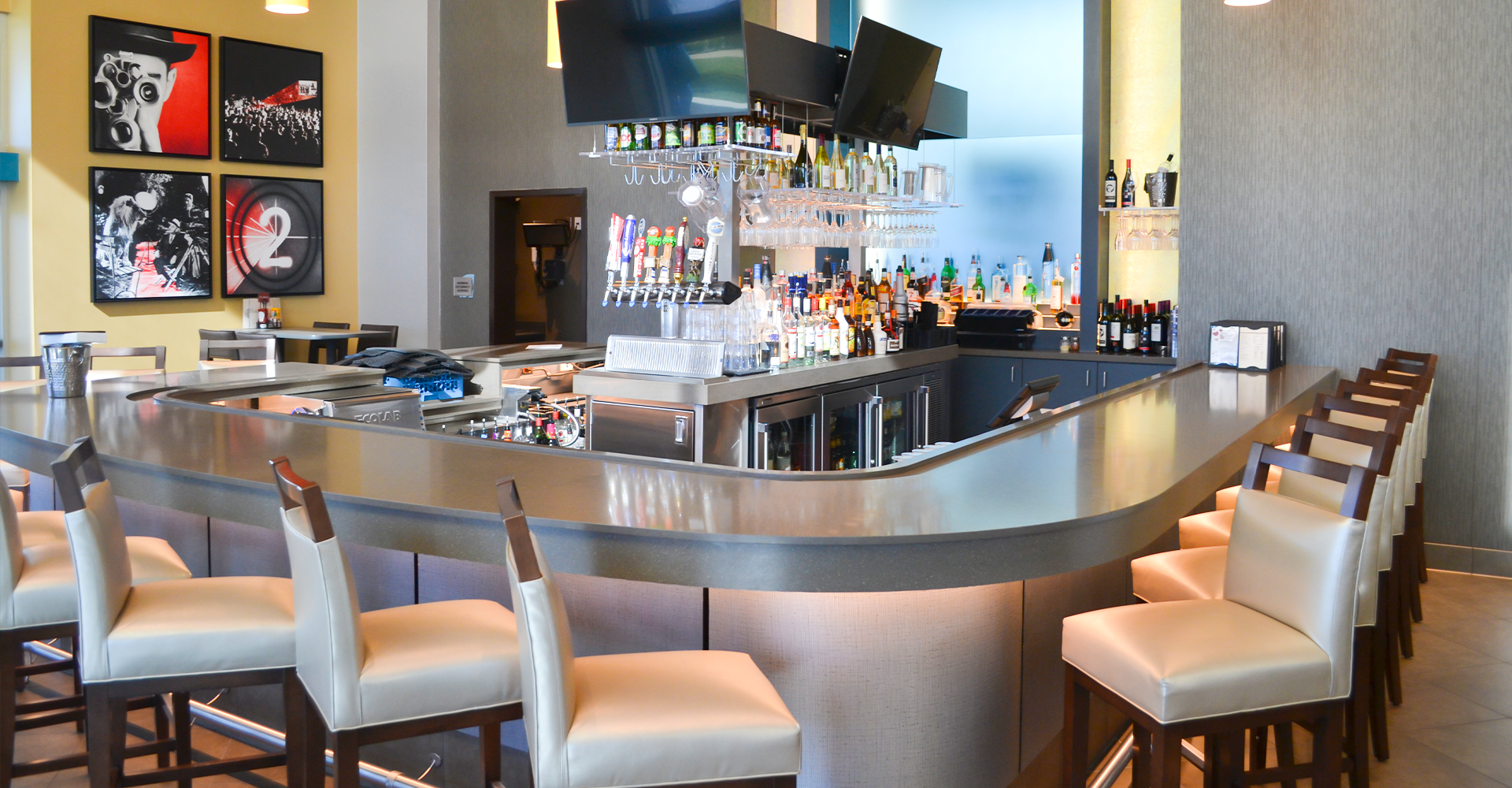 Bar with stainless steel counter, light-colored chairs, liquor bottles, and TVs. Artwork on the wall.