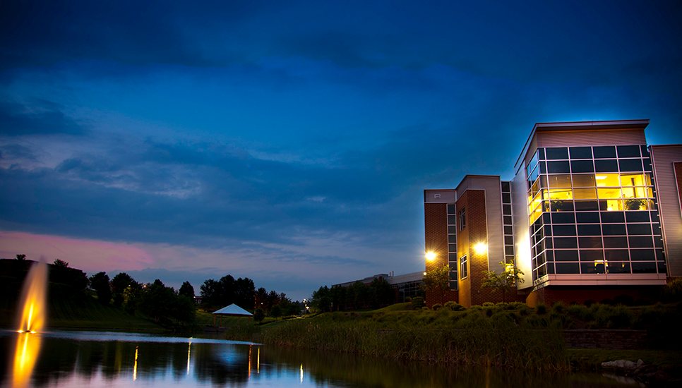 Modern building at dusk, illuminated windows reflect in the calm water with a fountain in the foreground.
