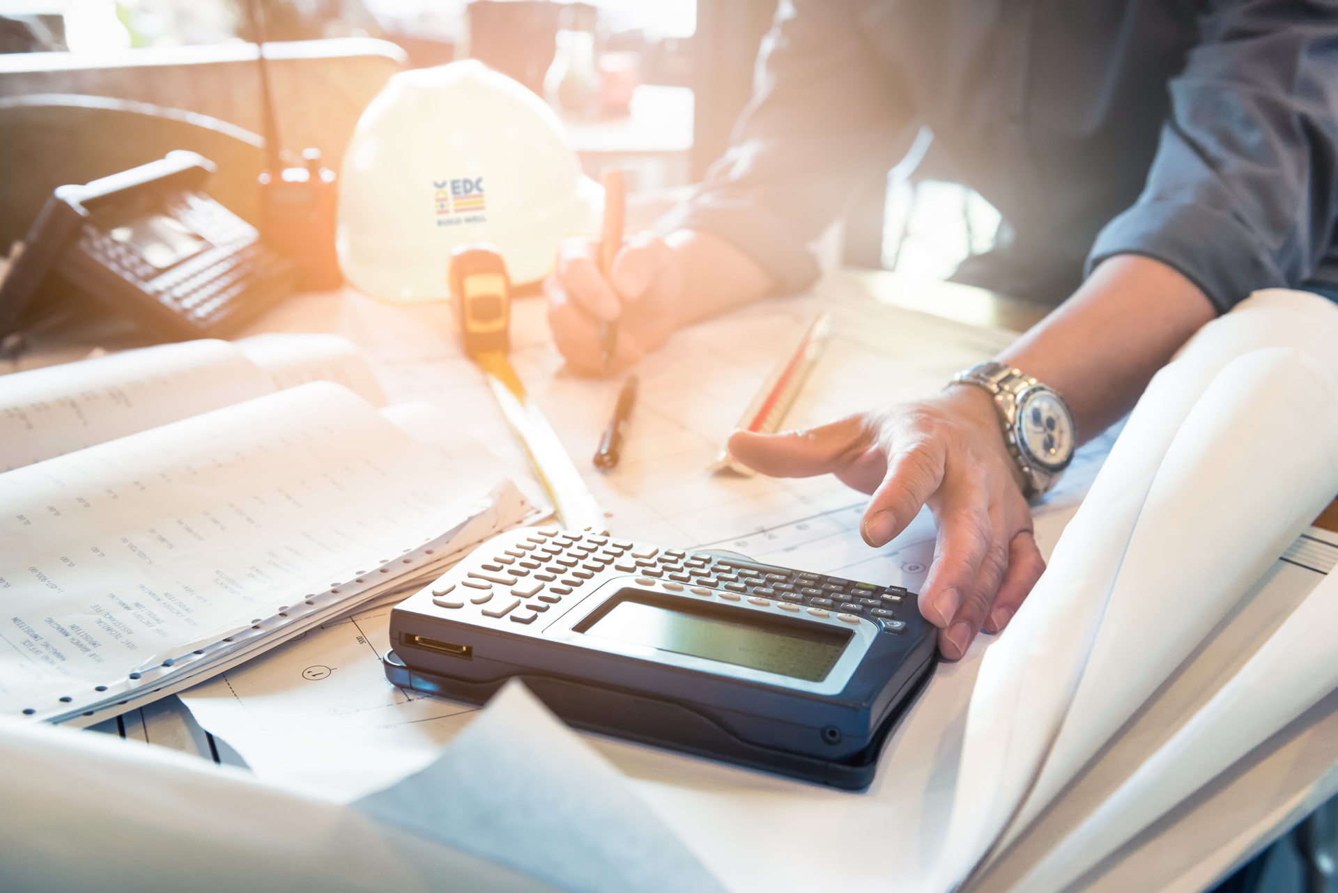 Person working on blueprints, using a calculator, with tools and a hard hat on a table.