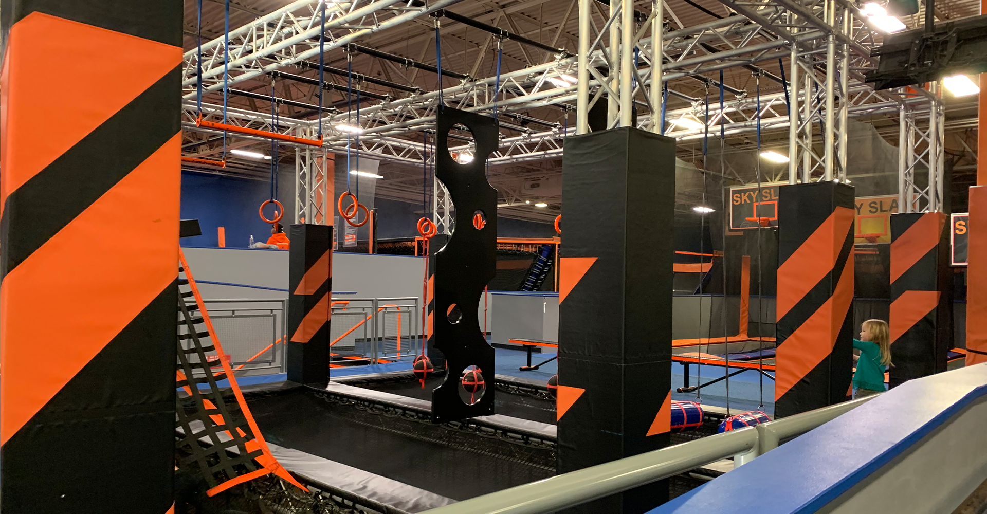 Indoor obstacle course, orange and black structures, hanging silhouettes, trampoline area.