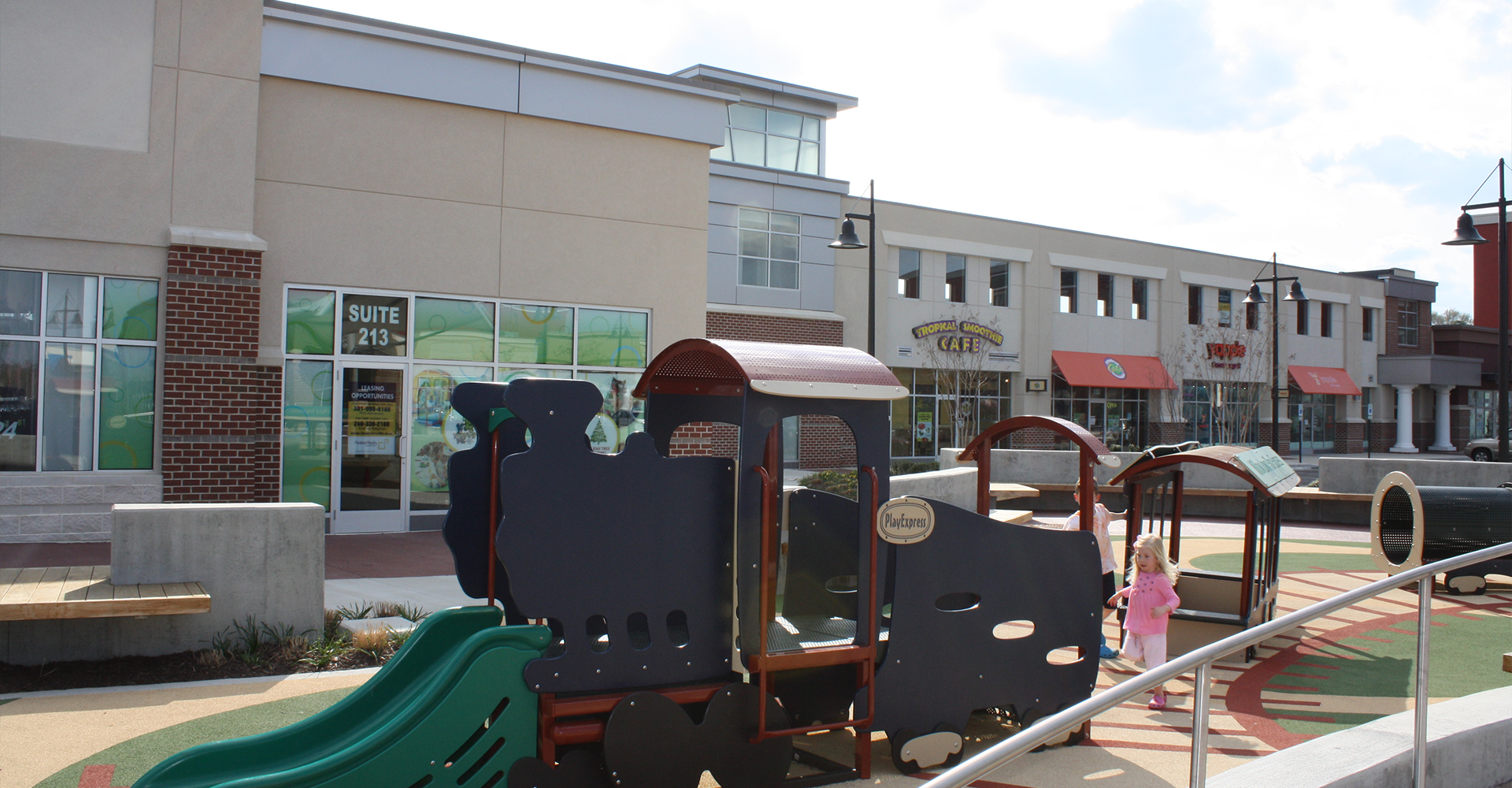 Playground with train structure in front of shops. A child in pink walks by.