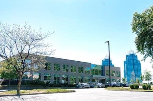 Modern office building with blue and gray facade, parking area, and tall blue skyscraper in the background.
