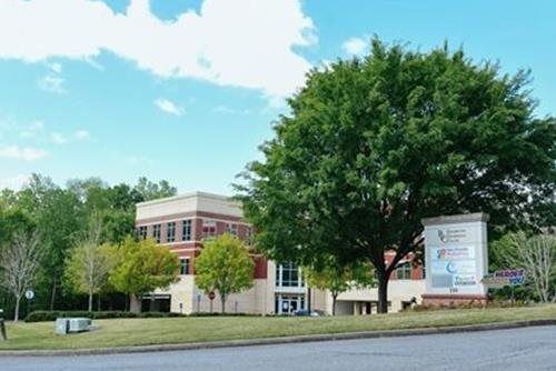 Office building with brick and beige facade, large tree, sign with text, blue sky.