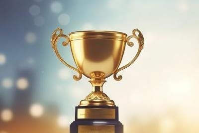 Gold trophy on a pedestal, with handles, against a blurred, light background.