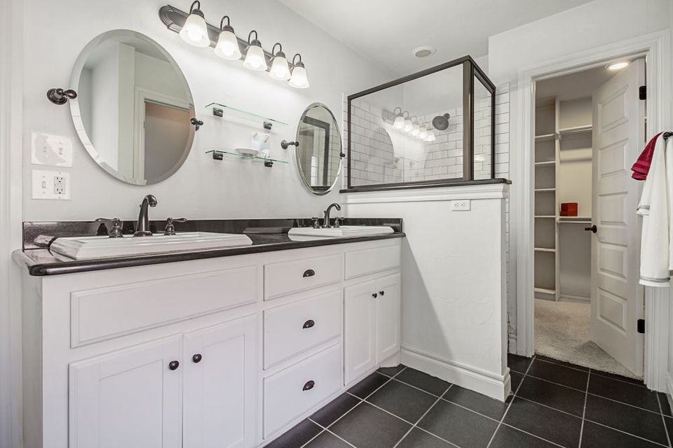 Modern white bathroom with black countertops, black floor tiles, and a walk-in shower. Double sinks with oval mirrors and a walk-in closet.