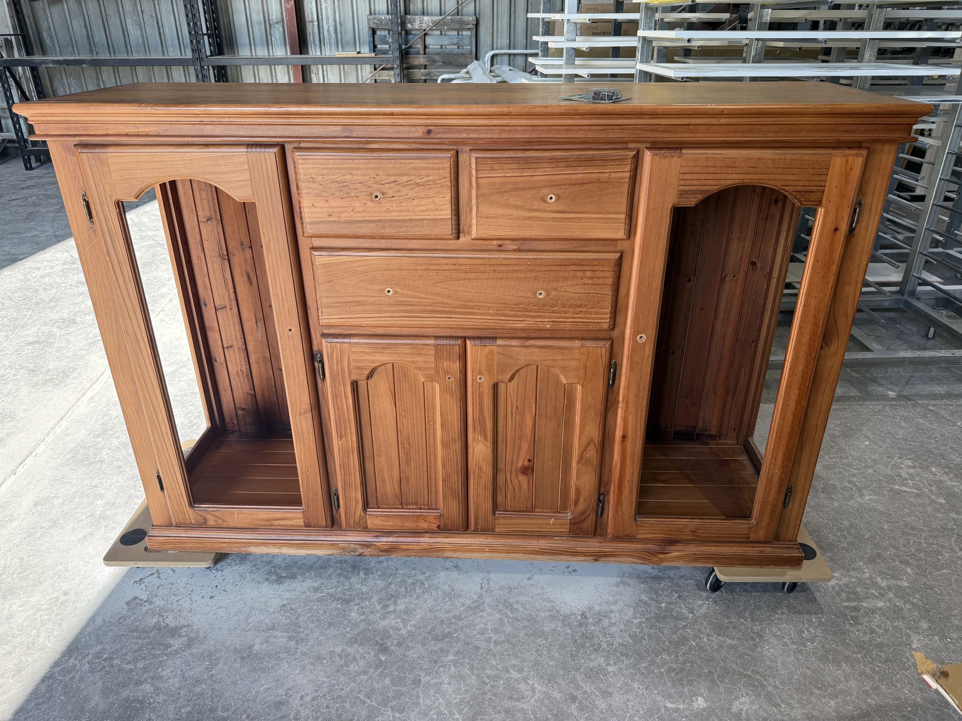 A wooden media console with two open side compartments, two small drawers, and two doors. It sits on a platform in a workshop.